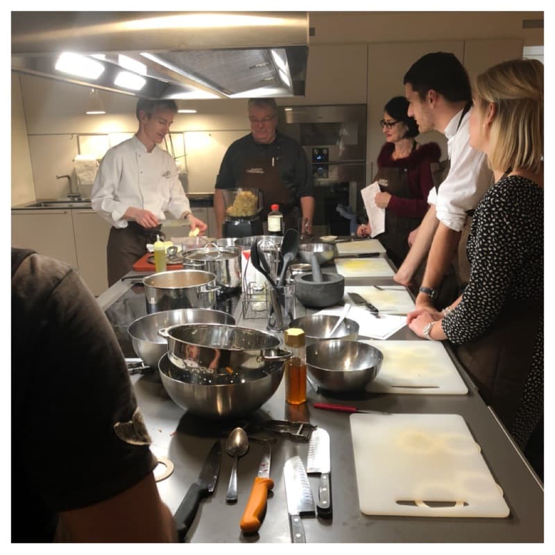 Un chef en toque blanche dirige un atelier culinaire autour d'un îlot de cuisine, plusieurs participants cuisinent ensemble avec enthousiasme