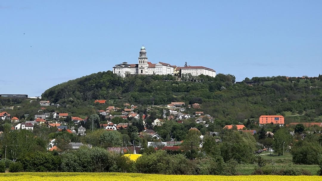 Château dominant une colline boisée avec un petit village aux toits rouges en contrebas, sous ciel bleu dégagé