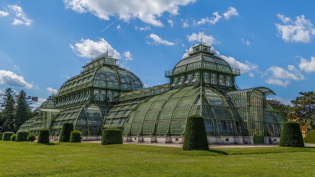 Palais de verre aux dômes arrondis et toiture verte entouré de pelouse et de sculptures de buis taillé, ciel bleu avec nuages