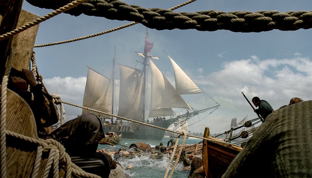Voilier de la série Black Sails avec voiles blanches et drapeau rouge, vue depuis le pont d'un navire pirate avec cordes et mâts au premier plan