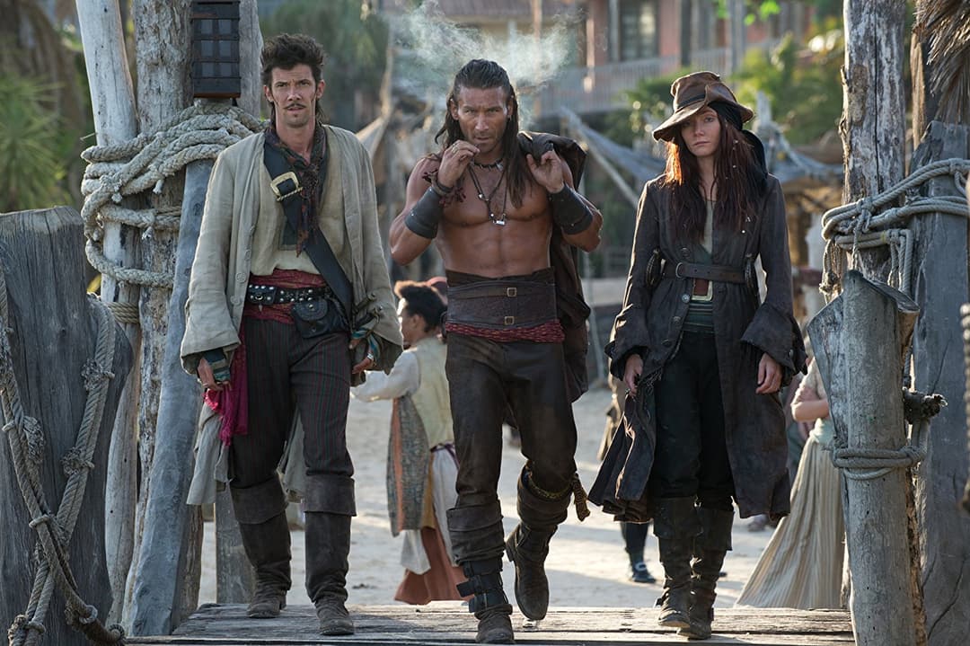 Trois pirates en costumes d'époque marchent dans une ruelle pavée d'une ville portuaire, encadrés par des bâtiments en ruine