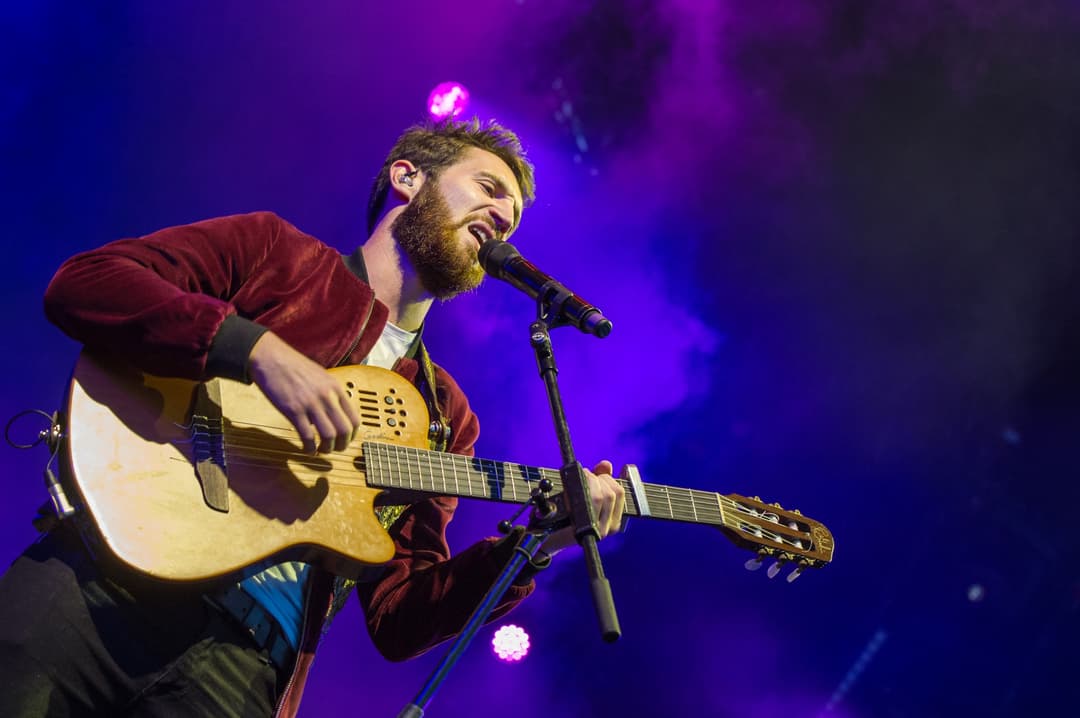 Musicien barbu chantant à la guitare acoustique sur scène, éclairage violet et bleu au Paléo Festival