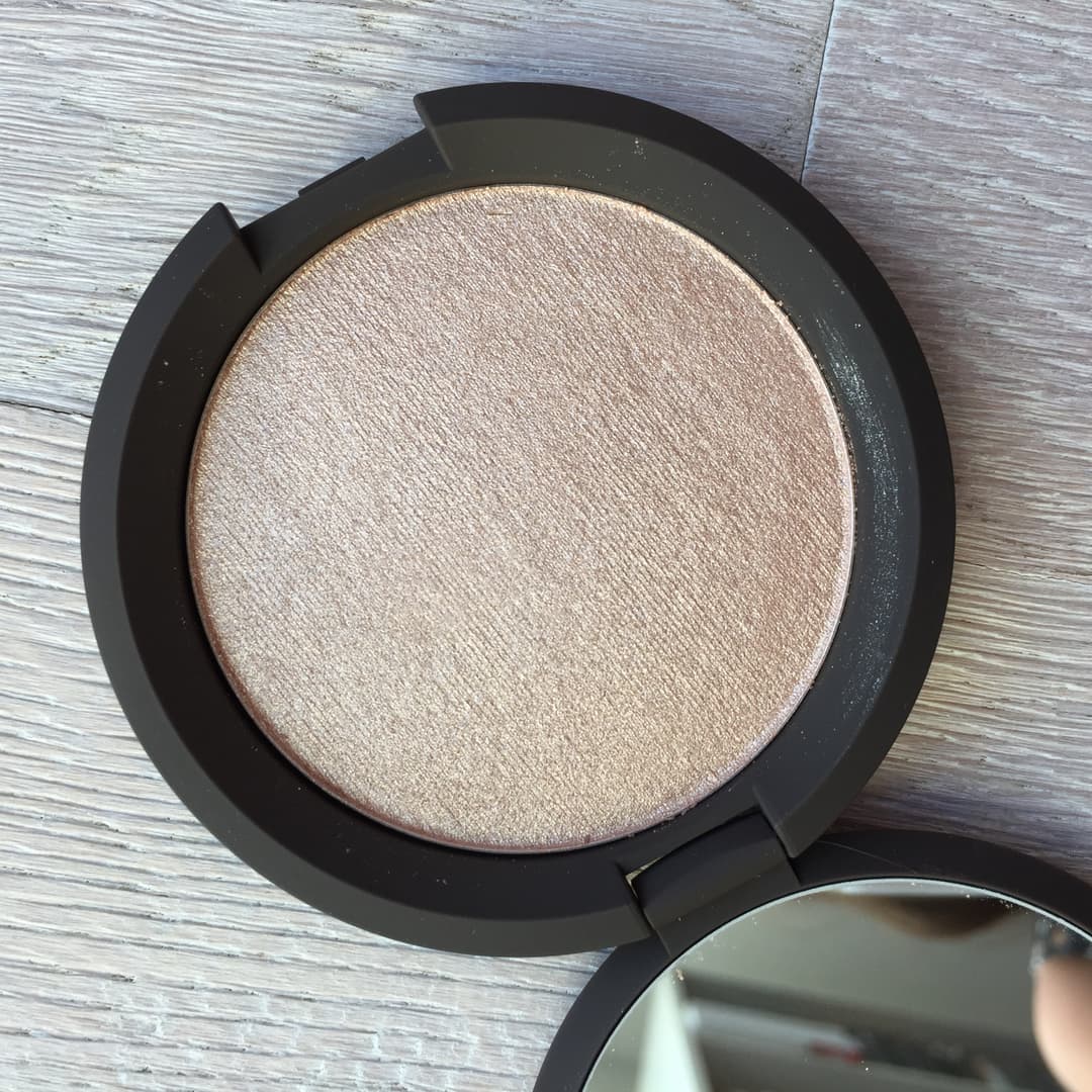 Becca Shimmering Skin Perfector, poudre beige nacré avec boîtier noir rond, sur surface bois gris clair