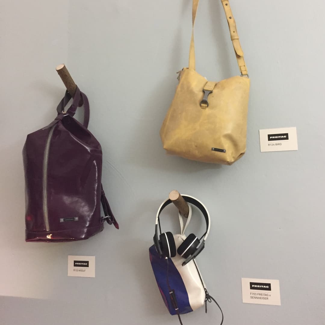 Trois produits Freitag : un sac à dos prune, un sac à bandoulière jaune paille et un casque noir et blanc posé sur un étui bleu