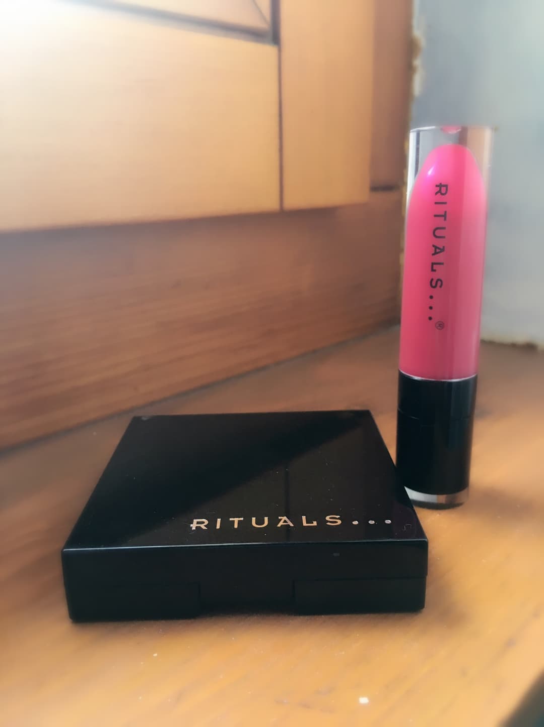Lipgloss rose vif Rituals et palette Magic Powder Blush noire de la marque, posés sur une table en bois