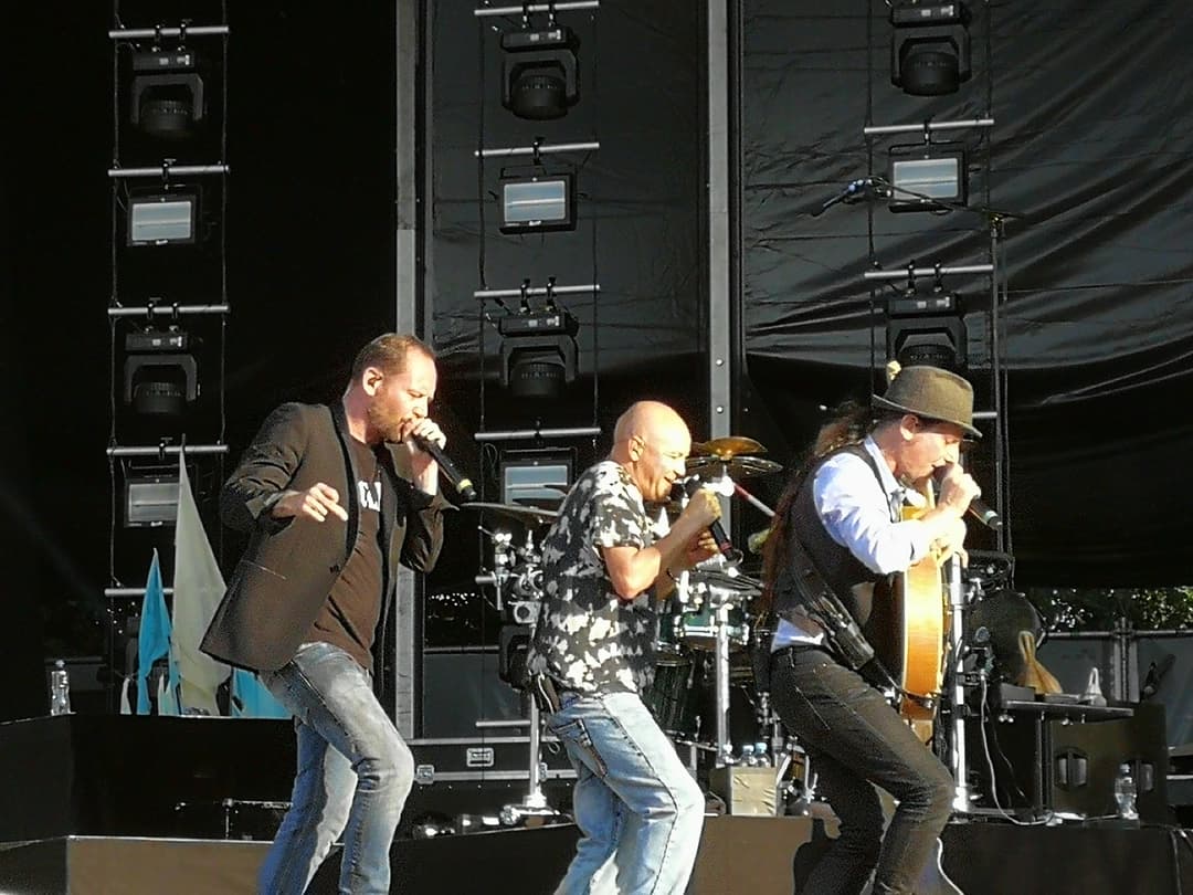 Trois chanteurs sur scène au Paléo Festival de Nyon, dont un à la guitare acoustique, devant un écran géant d'équipements techniques
