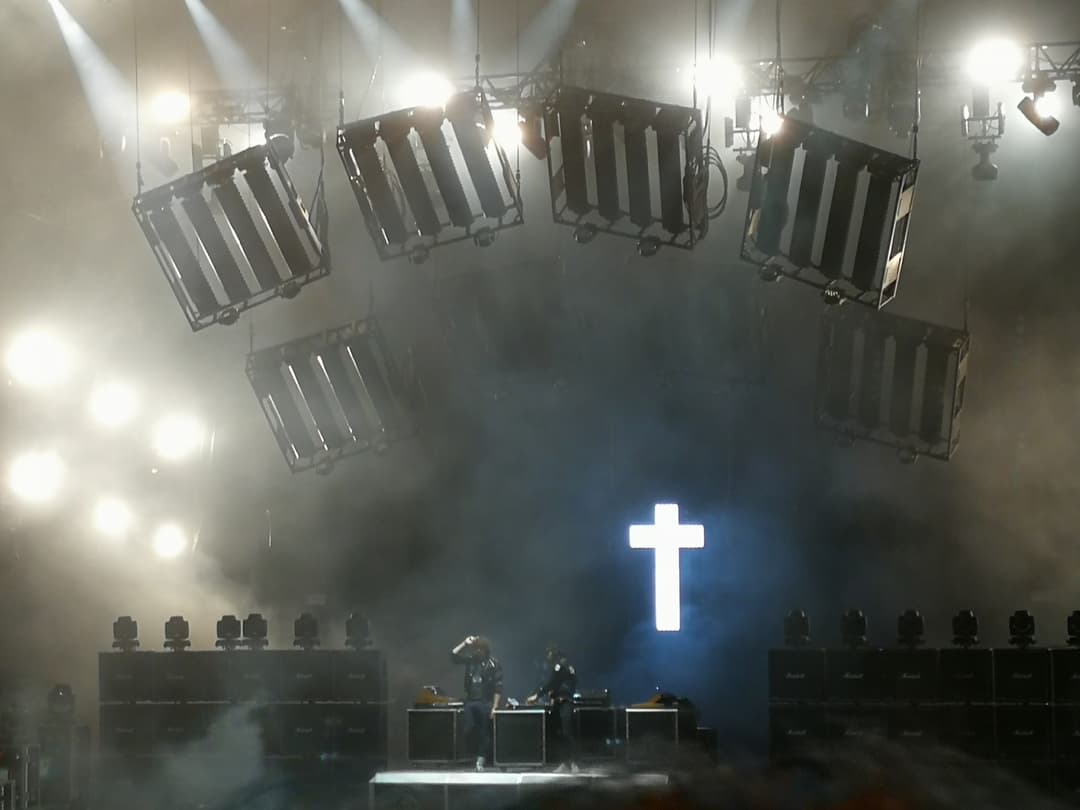 Scène du groupe JUSTICE au Paléo Festival, avec croix lumineuse bleue au centre et projecteurs suspendus au-dessus