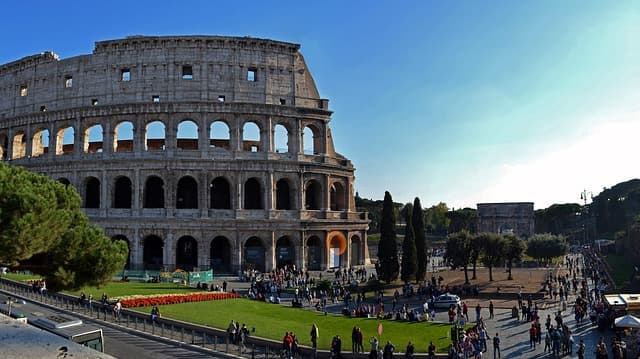 Le Colosseum de Rome avec ses arches monumentales, sous un ciel bleu, entouré de visiteurs sur une esplanade verte