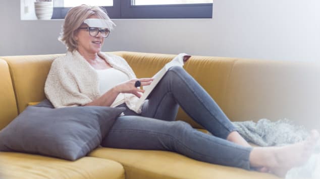 Femme portant les lunettes de luminothérapie Luminette, assise sur un canapé jaune, tenant une tablette