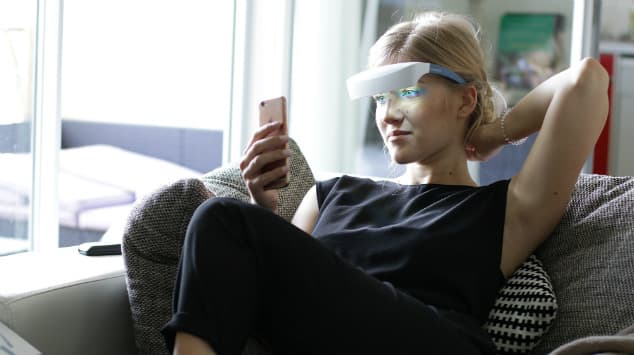 Femme portant des lunettes blanches de luminothérapie, assise sur un canapé, consultants son téléphone