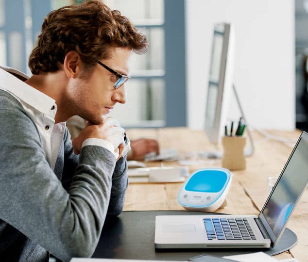 Homme avec lunettes travaillant à un bureau avec une lampe de luminothérapie Philips blanche et bleue devant son ordinateur portable