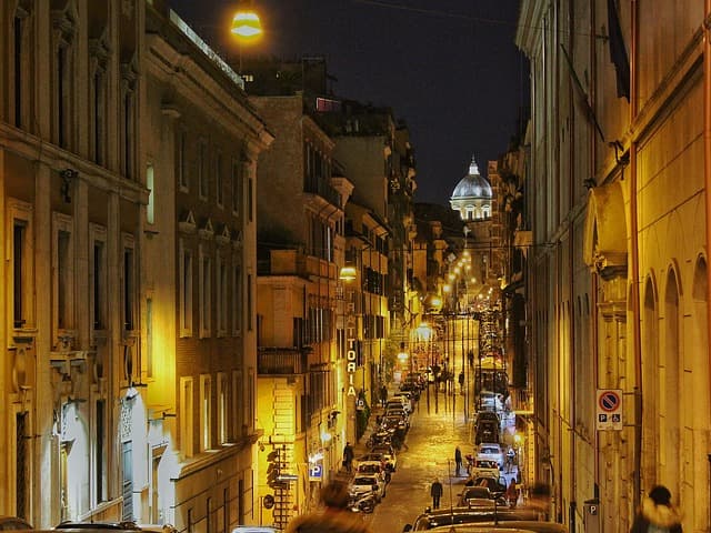 Rue étroite de Rome illuminée la nuit, avec des bâtiments historiques ocre de chaque côté et le dôme de Saint-Pierre en arrière-plan