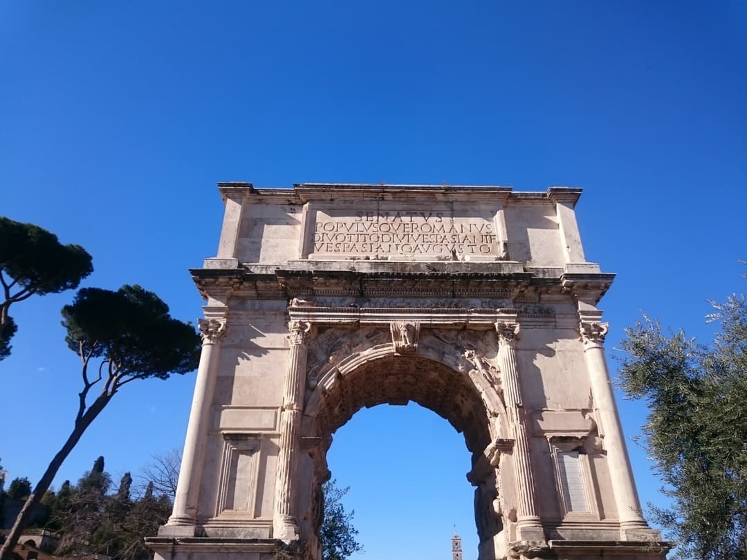 Arc de triomphe romain en marbre blanc avec inscription latine, arbres verts sur les côtés, ciel bleu dégagé