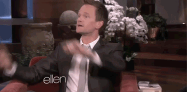 Un homme en chemise blanche et veste noire, assis sur un canapé, les mains levées, devant un logo Ellen