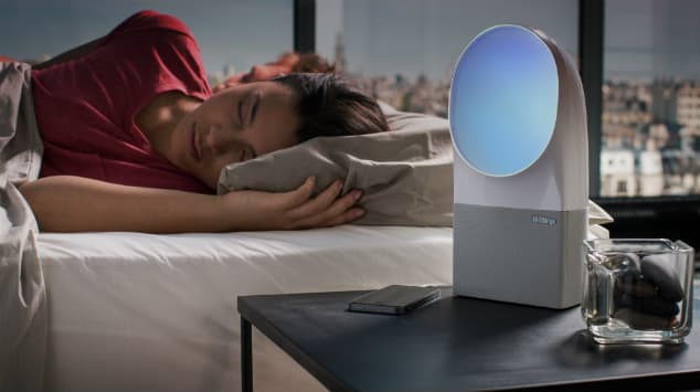 Appareil de luminothérapie blanc avec écran bleu rond sur table de nuit, à côté d'une personne endormie sous couverture rouge