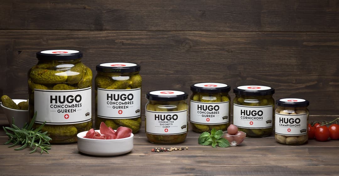 Coffret Hugo Reitzel composé de six pots en verre contenant des légumes marinés : concombres, cornichons et champignons, étiquetés HUGO, sur fond de bois