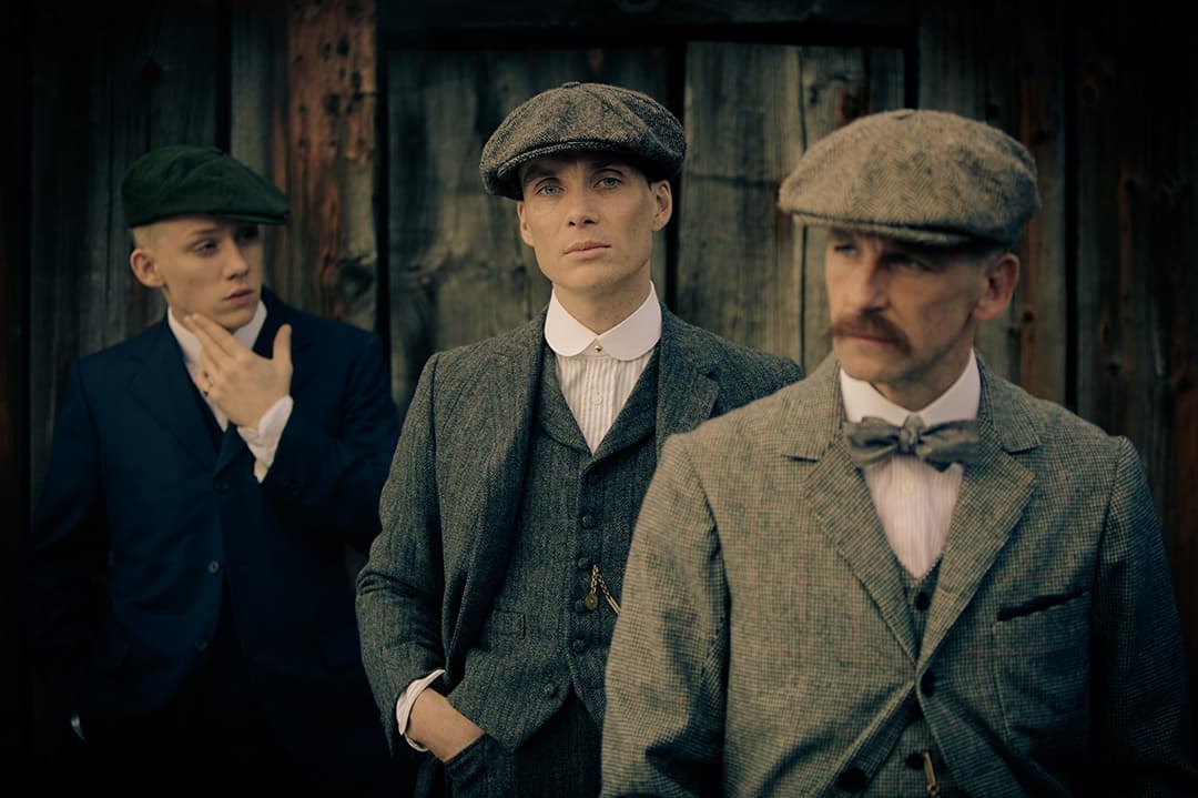 Trois hommes en costumes et casquettes d'époque années 1920 devant un bâtiment délabré, incarnant la famille Shelby de Peaky Blinders
