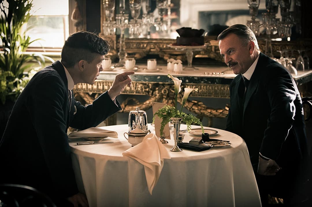 Deux hommes en costume noir discutent autour d'une table élégante dans un restaurant luxueux aux décors dorés et ornementés