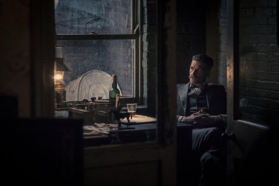 Personnage assis dans une pièce sombre aux murs de brique, éclairé par la lumière d'une fenêtre, avec une bouteille et un verre sur une table