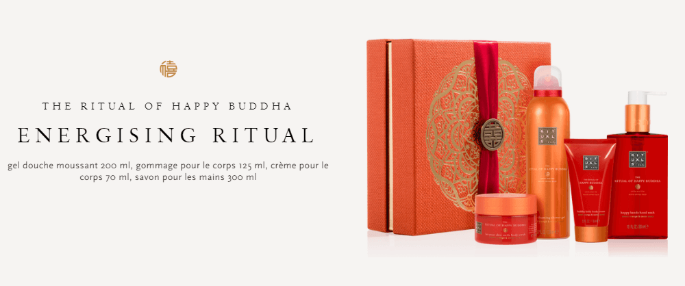 Coffret de soins The Ritual of Happy Buddha dans une boîte orange avec ruban rouge, contenant mousse douche, gommage, crème et gel lavant