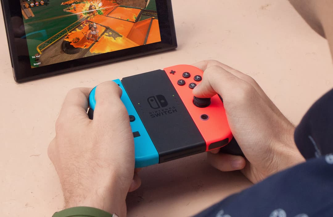 Nintendo Switch avec manettes bleue et rouge tenue en main, écran affichant un jeu en arrière-plan