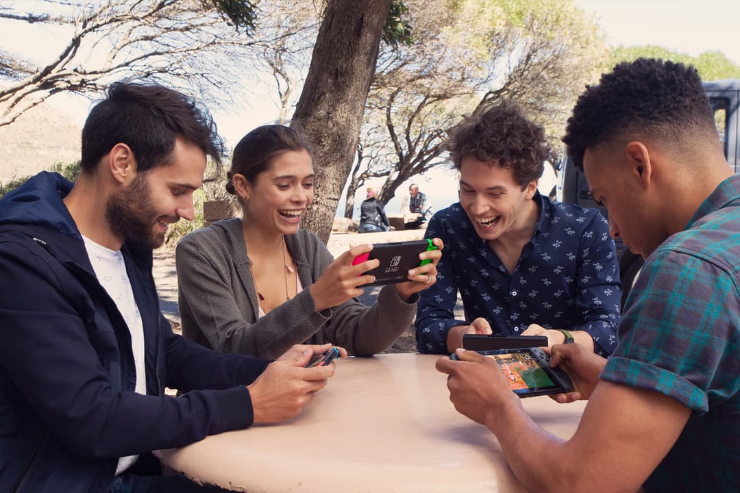 Quatre jeunes jouent ensemble à la Nintendo Switch en plein air, assis autour d'une table sous des arbres