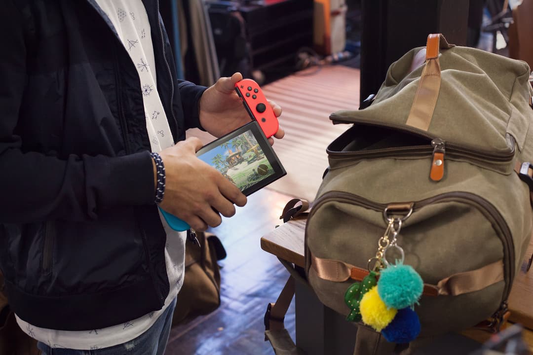 Nintendo Switch avec manette Joy-Con rouge tenue en main, écran affichant un jeu, à côté d'un sac à dos kaki avec porte-clés pompon