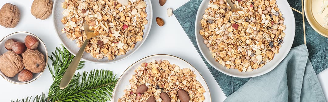 Trois bols de muesli avec fruits secs et noix, cuillère dorée, noix et brindilles vertes sur fond blanc