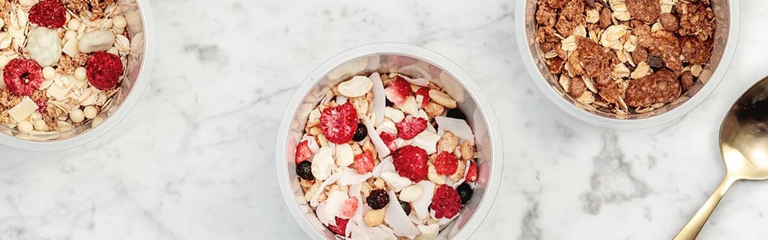Trois bols de céréales MYMUESLI contenant des flocons, des fruits rouges et des noix sur fond marbre blanc