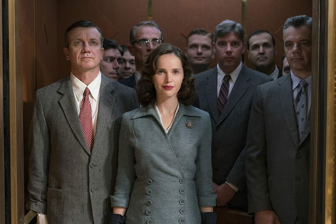 Ruth Bader Ginsburg entourée d'hommes en costumes dans un ascenseur, scène du film On the Basis of Sex