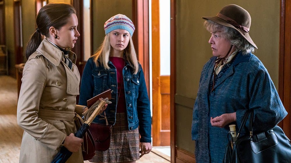 Trois femmes dans un intérieur rétro : une avocate en imperméable beige face à une jeune fille et une dame âgée en tenue vintage