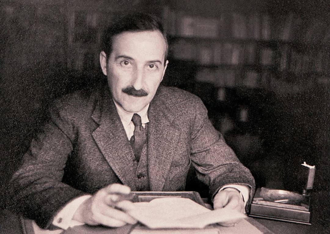 Portrait noir et blanc d'un homme à moustache en costume gris, assis à un bureau avec des feuilles et une cigarette
