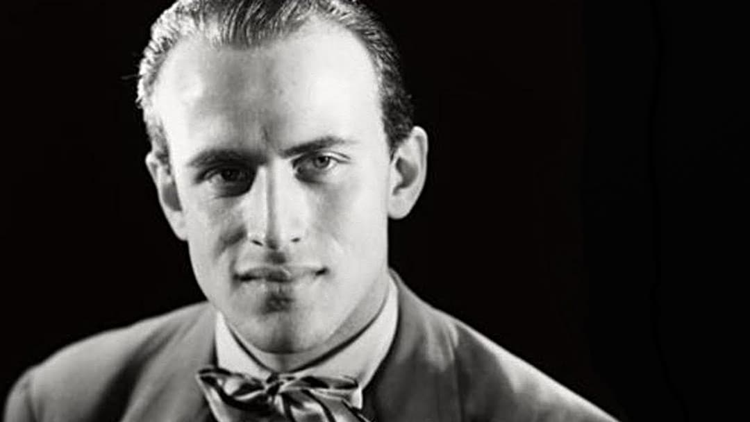 Portrait en noir et blanc d'un homme aux cheveux gominés, vêtu d'une chemise et d'un nœud papillon, photographié de face sur fond noir