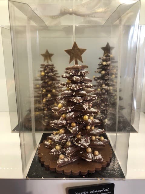 Sapin de Noël en chocolat noir garni de perles dorées et blanches, couronné d'une étoile, présenté dans une vitrine transparente avec décor hivernal en arrière-plan