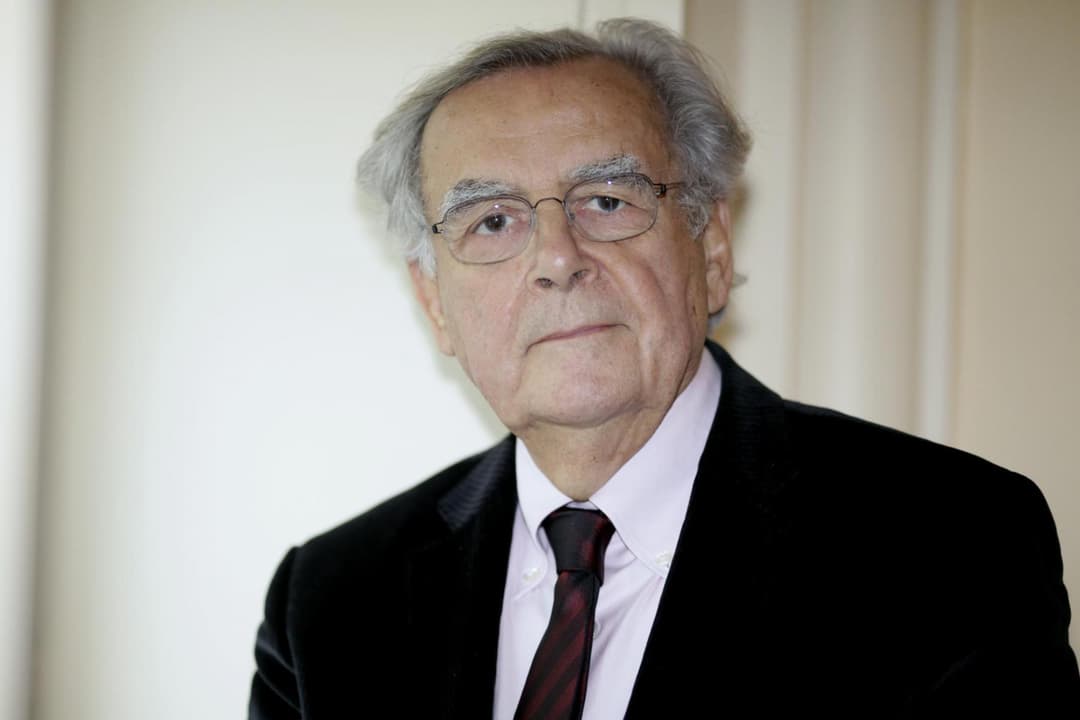 Homme âgé aux cheveux gris, portant des lunettes et un costume noir avec chemise rose et cravate bordeaux