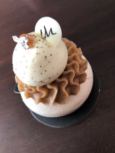 Pâtisserie meringué marron garnie de chantilly vanille, posée sur un socle noir, ornée d'une boule blanche et de feuille d'or