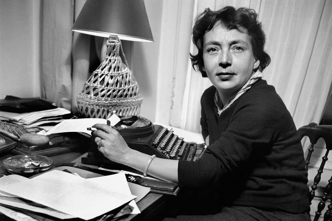 Femme écrivain assise à son bureau avec une machine à écrire, entourée de papiers et d'une lampe, en noir et blanc
