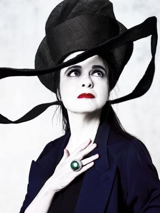 Femme au chapeau noir avec ruban, maquillage épuré et bague émeraude, vêtue d'un costume bleu marine