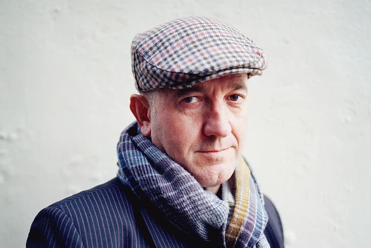 Homme portant une casquette à carreaux rose et bleu et une écharpe grise, vêtu d'une veste à rayures