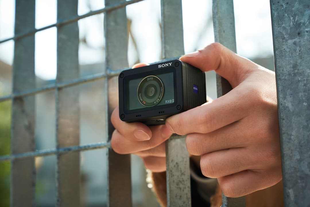 Sony RX0 II, appareil photo ultra-compact noir avec objectif ZEISS, tenu dans une main devant une clôture métallique