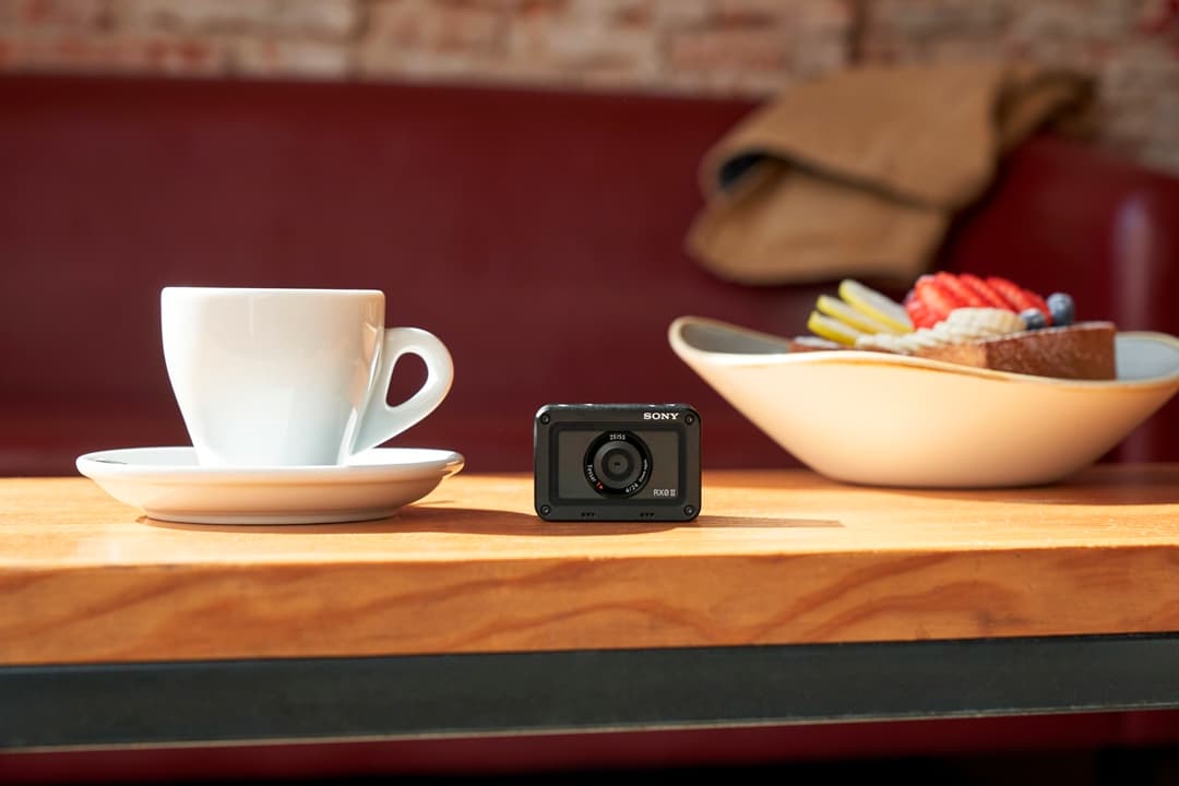 Sony RX0 II, petit appareil photo compact noir, posé sur une table en bois entre une tasse de café blanche et un bol de fruits