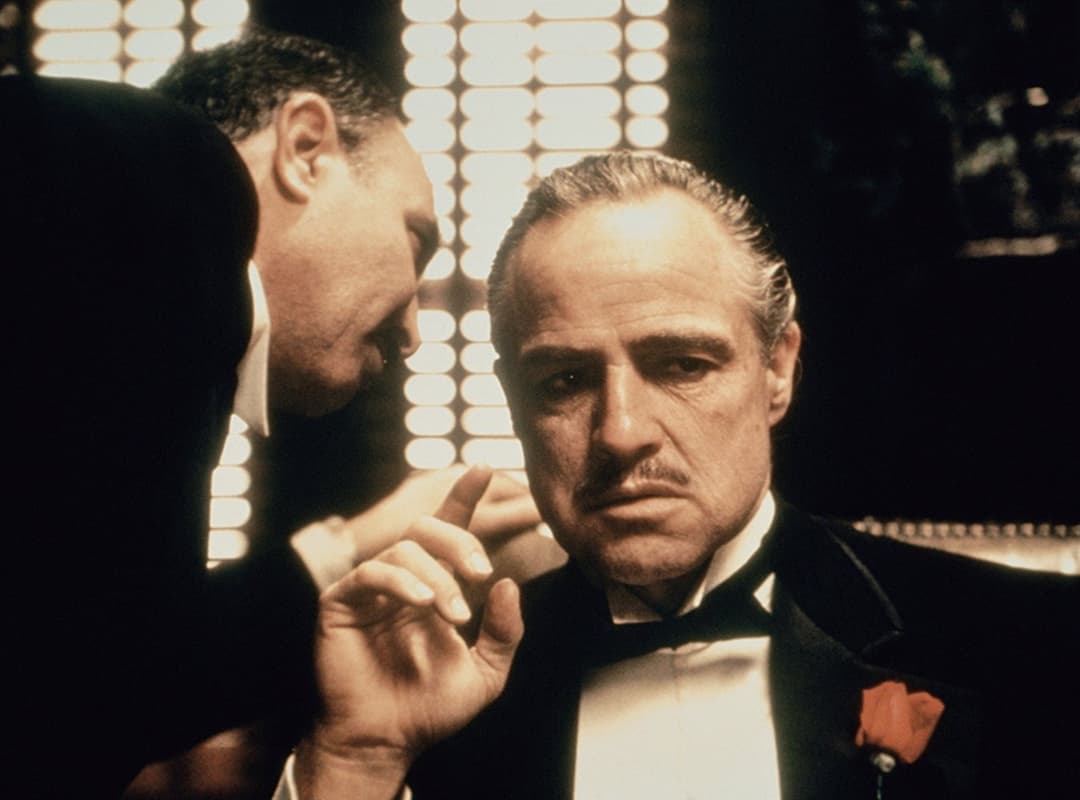 Deux hommes en costume et nœud papillon lors d'une conversation formelle, l'un de profil, l'autre de face, dans une salle richement décorée
