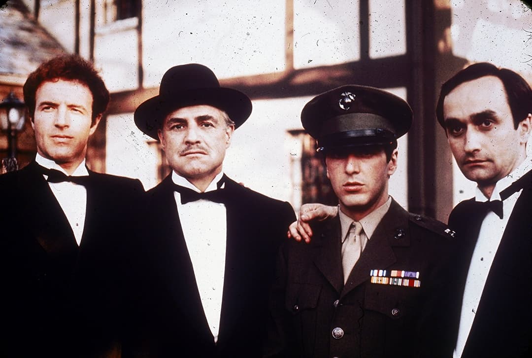 Quatre hommes en costumes sombres et chapeaux, dont un en uniforme militaire, dans une lumière contrastée typique du Parrain