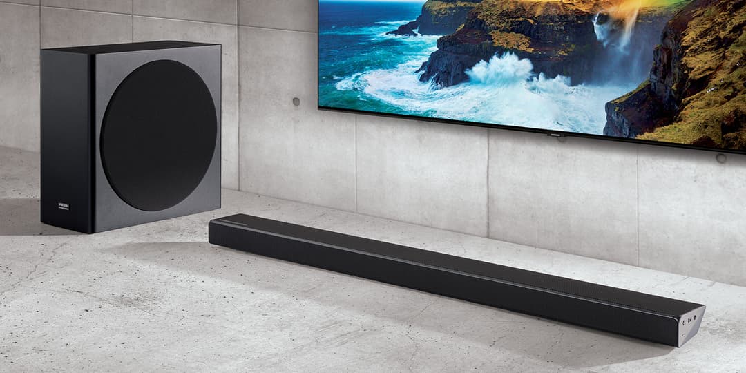 Barre de son Samsung série Q noire allongée au sol, accompagnée d'un caisson de basses circulaire, face à un téléviseur QLED affichant une falaise côtière