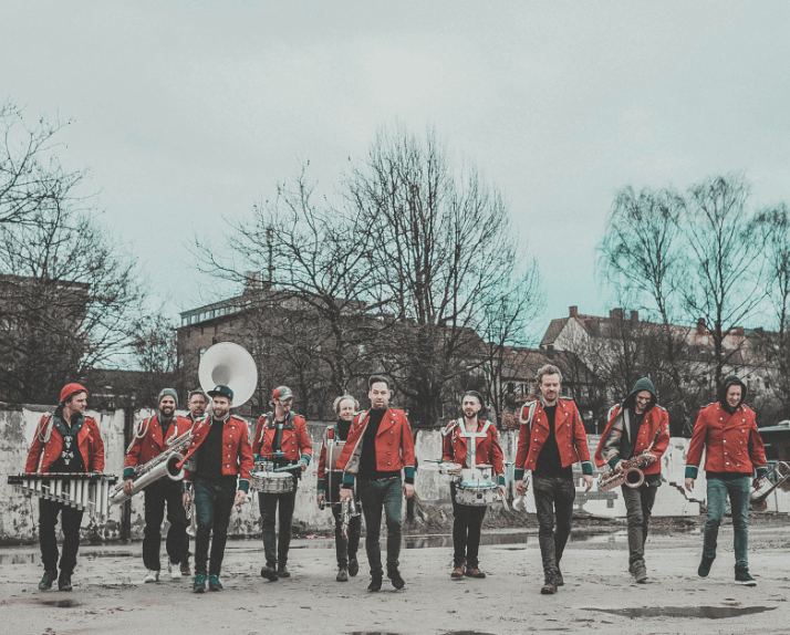 Groupe de musiciens en vestes rouges jouant des instruments à vent et percussion dans une cour urbaine avec immeubles en arrière-plan