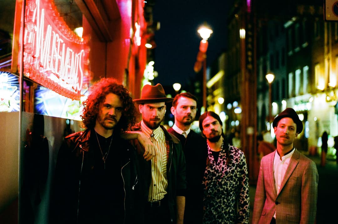 Groupe de cinq musiciens posant dans une rue illuminée de néons rouges et jaunes la nuit