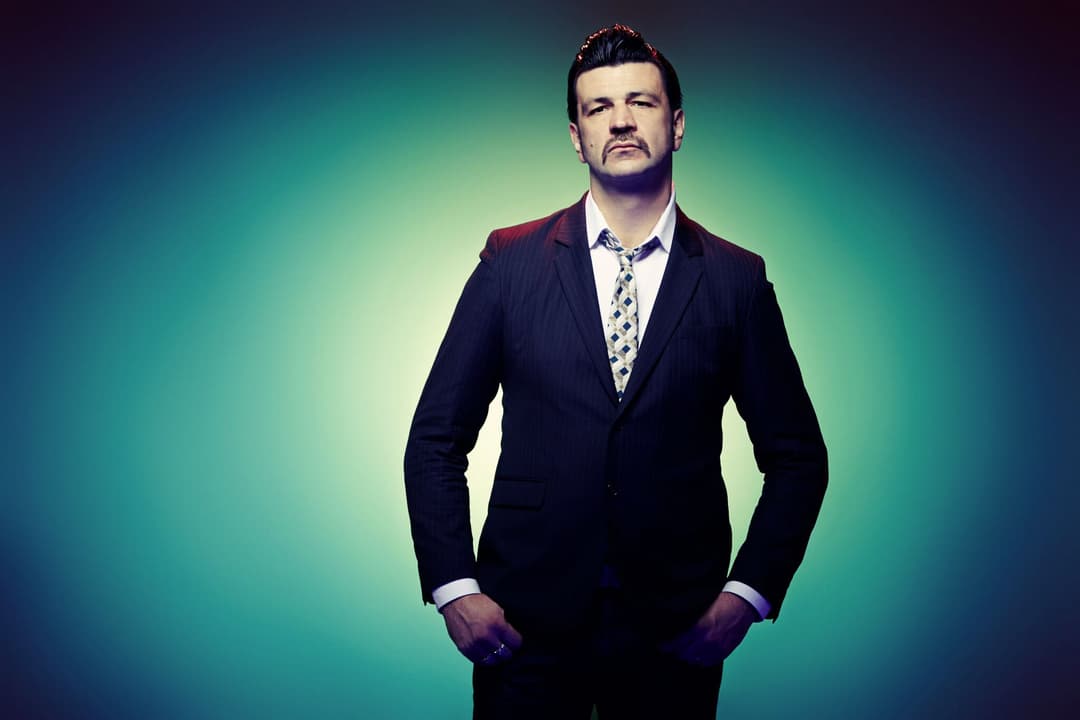 Arnaud Rebotini en costume noir et chemise à motifs, les mains dans les poches, sur fond dégradé bleu et vert