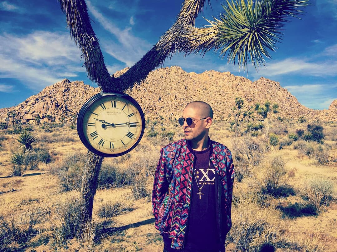 Arnaud Rebotini en lunettes de soleil et veste colorée posant devant une horloge dans un désert rocailleux
