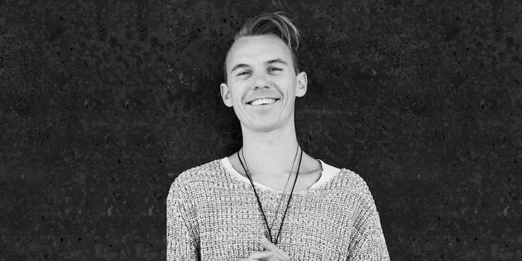 Portrait en noir et blanc d'un jeune homme souriant portant un badge autour du cou, devant un mur sombre