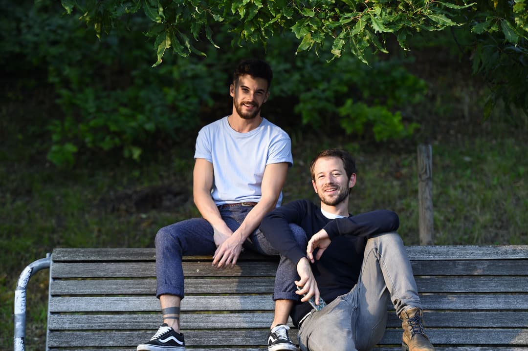 Kyrill & Redford assis sur un banc en bois devant un mur de verdure, souriant à la caméra